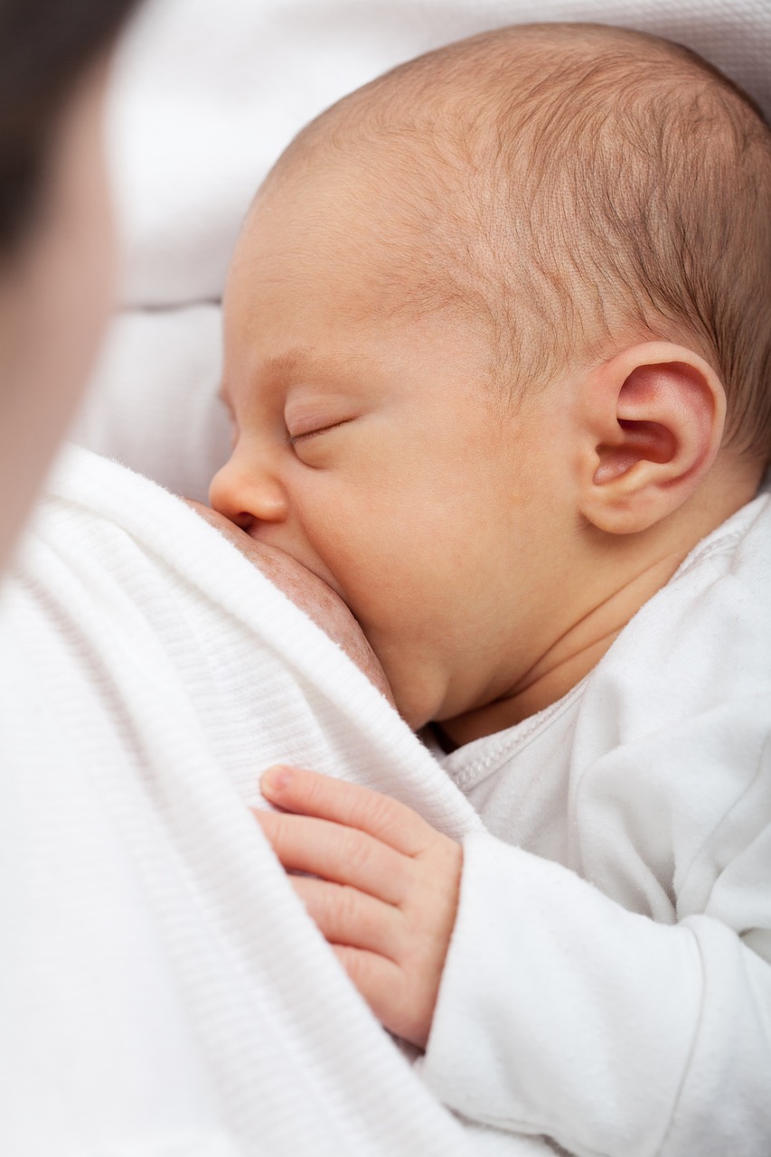 Ein Neugeborenes in weißer Kleidung wird von seiner Mutter gestillt. Der Säugling ist ruhig und schläft möglicherweise, während es die Brust der Mutter hält und daran saugt. Dieser Beitrag bietet 12 Stilltipps für Mütter, um das Stillen erfolgreich und stressfrei zu gestalten.